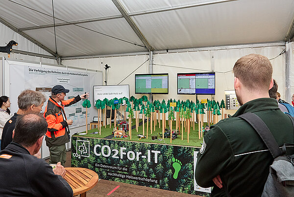 Messestand mit Menschen um einen Modellbau-Wald auf einem Tisch und Monitore